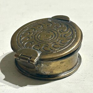 Puede incluir: Una pequeña pastillera redonda de latón con tapa abatible. La parte superior de la tapa presenta un patrón circular en relieve con motivos curvos repetitivos y un punto central, rodeado por un borde texturizado. El metal tiene un acabado patinado.