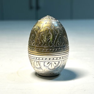 Może przedstawiać: Metalowy przedmiot w kształcie jajka o dwukolorowym wykończeniu, z górną częścią w kolorze złotym i dolną w kolorze srebrnym. Powierzchnia jest ozdobiona misternymi, grawerowanymi wzorami geometrycznymi oraz stylizowanymi motywami liniowymi na całej powierzchni.