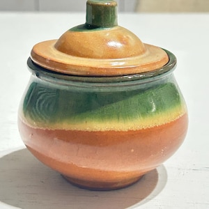 Può includere: Un piccolo barattolo in ceramica rotondo con coperchio abbinato, caratterizzato da una smaltatura lucida a bande orizzontali nei colori arancione terracotta, verde erba vivace e giallo oro caldo. Il coperchio presenta un piccolo pomello.