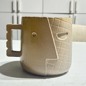 Op de afbeelding: Een beige keramische mok met een gladde afwerking aan de ene kant en een rasterpatroon aan de andere kant. De mok heeft een gestileerd gezichtsontwerp met één cirkelvormig oog en een geometrisch handvat met een gekartelde buitenrand.
