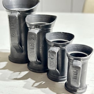 Puede incluir: Un juego de cuatro jarras medidoras graduadas de metal gris oscuro con asas. Cada jarra presenta texto en relieve en el asa que indica su capacidad: 1 DECILITER, 1/2 DECILITER, 2 CENTILITRES y 1 CENTILITRE, ordenadas de mayor a menor tamaño.