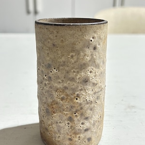 May include: A cylindrical ceramic tumbler featuring a textured, mottled surface with a beige and tan colour palette. The exterior displays a porous, cratered finish with subtle dark brown speckles and a smooth, dark rim along the top edge.
