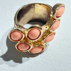 Puede incluir: Un anillo de tono dorado que presenta un grupo de cabujones lisos, redondeados y de color melocotón engastados en una banda de metal texturizada. El diseño incorpora múltiples engastes circulares de varios tamaños que envuelven el dedo, creando una apariencia audaz y orgánica con un acabado pulido.