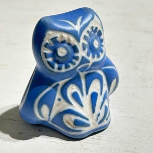 Puede incluir: Una pequeña figura de búho con un esmalte azul vibrante y patrones incisos en blanco que contrastan. El diseño incluye ojos circulares con líneas radiantes y un motivo de plumas estilizado en el pecho, creando una apariencia bicolor.