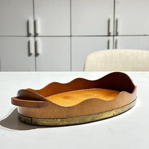 Pode incluir: Uma bandeja oval de madeira com uma borda ondulada e festonada e duas pequenas alças laterais. A base da bandeja é cercada por uma faixa de metal de latão desgastado e manchado que contorna o perímetro inferior, contrastando com a madeira.