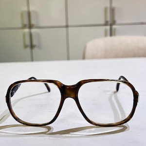 Könnte beinhalten: Brille mit einem dicken, rechteckigen Rahmen in braun-schwarzer Schildpatt-Optik. Die Brille hat klare Gläser und dünne, dunkel gefärbte Bügel, die von den Seiten des Rahmens ausgehen und einen klassischen, strukturierten Look erzeugen.
