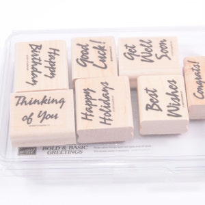 Puede incluir: Un juego de ocho sellos de goma de madera con varios saludos, que incluyen "Feliz cumpleaños", "Buena suerte", "Que te mejores pronto", "Pensando en ti", "Felices fiestas", "Los mejores deseos" y "¡Felicidades!" Los sellos están en una caja de plástico transparente.