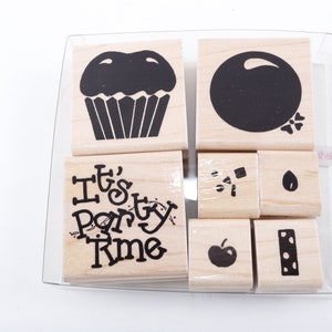 Puede incluir: Un juego de sellos de madera con diseños de tinta negra, que incluyen una magdalena, un círculo y la frase "It's Party Time". Otros sellos presentan una manzana, una semilla y formas abstractas. Los sellos están en un recipiente de plástico transparente.