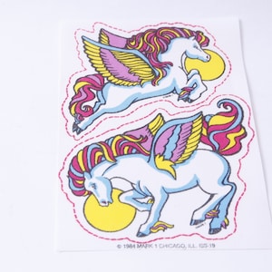 Op de afbeelding: Twee kleurrijke gevleugelde paardenstickers met roze, gele en blauwe details. De stickers bevinden zich op een witte achtergrond met een copyrightvermelding onderaan: © 1984 MARK 1 CHICAGO, ILL 1SS-19.