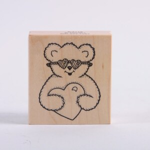 May include: A wooden rubber stamp featuring a black and white illustration of a teddy bear wearing sunglasses and holding a heart.