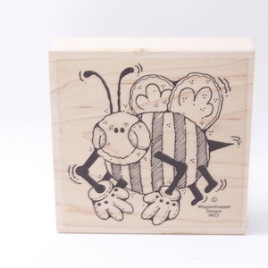 May include: A wooden stamp featuring a black and white illustration of a cartoon bee with a striped body and large eyes. The bee is wearing gloves and has a happy expression. The stamp is labelled "WhipperSnapper Designs" and "M115".