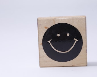 May include: A wooden block stamp with a black smiley face design. The smiley face has white eyes and a white smile.