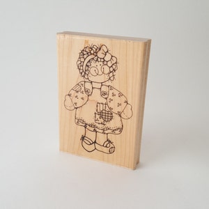 Puede incluir: Un bloque de madera con un dibujo en blanco y negro de una muñeca de trapo con un lazo en el pelo. La muñeca lleva un vestido con un bolsillo en forma de corazón.