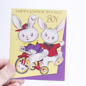 May include: A vintage Easter card with two white bunnies. One bunny is riding a red tricycle and the other bunny is standing behind it. The card has a purple border and the text "Happy Easter to a nice boy" is printed on the front.