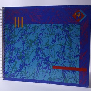 May include: A blue spiral bound notebook with a marbled design in shades of blue and green. The cover has a red, yellow, and blue geometric design in the top right corner.