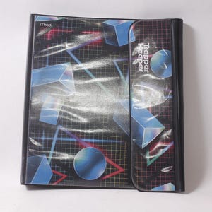 May include: A black Mead Trapper Keeper binder featuring a grid pattern background with blue geometric shapes, including cubes and spheres. The front flap displays the Trapper Keeper logo in white text, and the design includes red triangle accents.
