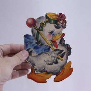May include: A vintage birthday card featuring a cartoon duck wearing a green hat with a red feather and holding a yellow stick with a red ball on top. The card says "Happy Birthday" in blue lettering.