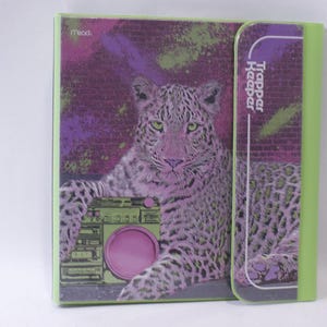May include: A lime green Trapper Keeper binder featuring a graphic of a leopard with purple and green tones. The leopard is positioned next to a green boombox. The text Trapper Keeper is printed vertically on the purple closure flap.