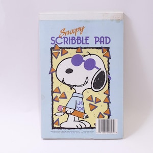 May include: A light blue Snoopy Scribble Pad featuring Snoopy wearing purple sunglasses and holding a pencil. The pad has the words "Snoopy Scribble Pad" in orange and a yellow border with orange triangles.