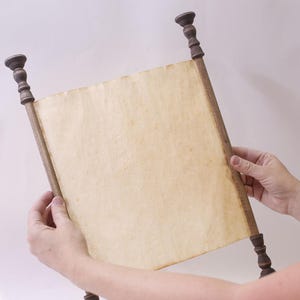 Puede incluir: Un pergamino de estilo vintage con papel envejecido, sostenido por manos. El pergamino está enmarcado por espigas y remates de madera marrón oscuro. El papel tiene un aspecto amarillento y texturizado, lo que sugiere edad o calidad de pergamino.