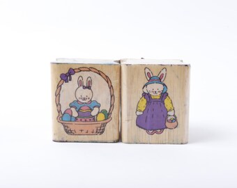 Può includere: Due timbri in legno con disegni di coniglietti di Pasqua. Un timbro presenta un coniglietto che tiene un uovo di Pasqua in un cestino. L'altro timbro presenta un coniglietto che tiene un cestino di uova di Pasqua.