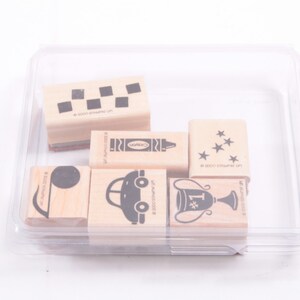 May include: A set of six wooden stamps in a clear plastic container. The stamps feature various designs, including a car, a trophy, a crayon, a baseball, stars, and a checkered pattern.