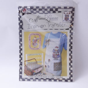 Könnte beinhalten: Eine Packung mit einem Bügelbild für eine Schürze mit einem Comic-Engel, der einen Blumenkorb hält. Die Packung ist gelb mit einem schwarz-weiß karierten Rand. Der Text auf der Packung lautet "Alma Lynne Originals Iron-on Transfer".