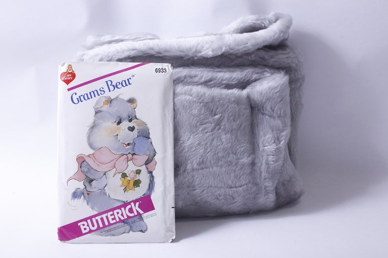 May include: A vintage Care Bears sewing pattern featuring Grams Bear, a blue bear with a pink ribbon and a yellow rose. The pattern is from Butterick and has the number 6933.