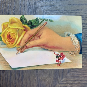 May include: A vintage postcard featuring a hand writing on a blank sheet of paper with a yellow rose and a small sprig of red flowers. The hand is holding a gold pen.