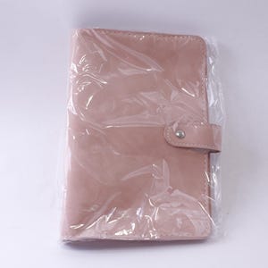 May include: A blush-colored, leather-like planner or notebook, sealed in clear plastic. The cover has a rounded flap with a snap closure. The planner is rectangular and appears to be new.