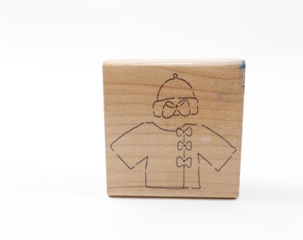 May include: A wooden stamp featuring a line drawing of a person wearing a hat and a sweater with bows.