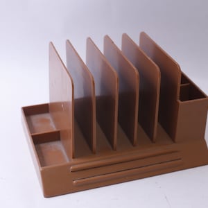 May include: A brown plastic desk organiser with six vertical compartments and a small drawer. The organiser has a textured surface.