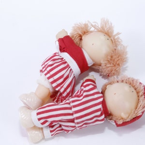 Puede incluir: Dos muñecas de tela a rayas rojas y blancas con pelo de peluche. Una muñeca lleva una camisa roja y la otra una chaqueta con capucha roja.