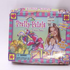 Könnte beinhalten: Eine "Pretty Petals Flower Maker"-Box mit einem Mädchen, das Blumen hält. Die Box zeigt Blumenillustrationen und den Text "Just Paint 'N Peel Petals!" und "Make Shimmering Flowers". Die Box ist mehrfarbig.