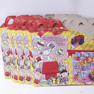 May include: A stack of eight Peanuts-themed Happy Meal boxes from McDonald's. The boxes feature a red and yellow design with images of Snoopy, Charlie Brown, and other Peanuts characters. The boxes also include a game where you match the wishes to the characters who made them. The back of the boxes feature a Tonka truck game.