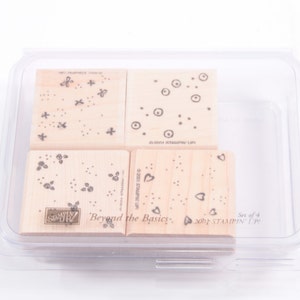 Peut inclure: Un ensemble de quatre tampons en bois avec différents motifs, y compris des points, des cœurs et des étoiles. Les tampons sont emballés dans un étui en plastique transparent et étiquetés "Beyond the Basics" et "Stampin' Up!"