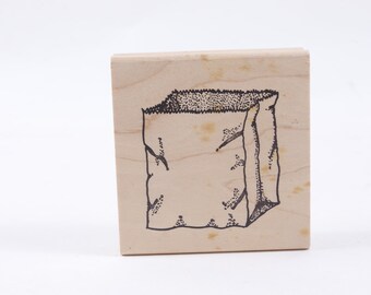 May include: A wooden stamp with a black and white illustration of a paper bag.