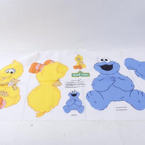 May include: A white fabric panel featuring Sesame Street characters. Big Bird is shown in yellow with orange feet and a teddy bear. Cookie Monster is blue, and there is a small yellow bird and a blue Cookie Monster. The Sesame Street logo is also visible.