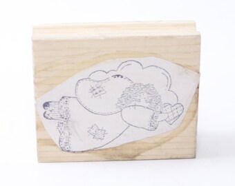 May include: A wooden stamp with a design of a cartoon angel holding a heart. The angel is wearing a dress with a patched design.