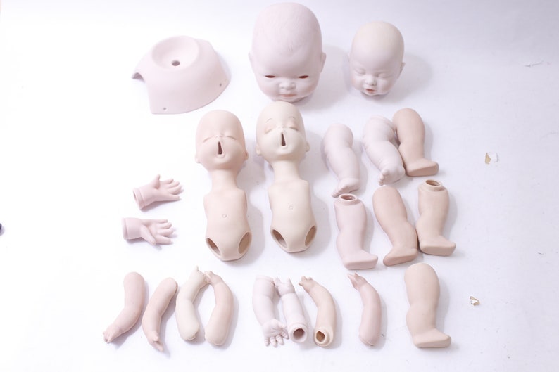 May include: A set of unpainted porcelain doll parts, including two heads, two torsos, two pairs of arms, four legs, and a head base. The doll parts are all white and have a smooth, matte finish.
