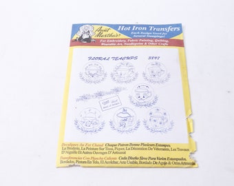 May include: A white sheet of paper with a yellow border. The paper has a blue and red illustration of a woman in a dress and a white apron. The text "Aunt Martha's Hot Iron Transfers" is printed in black. The text "Each Design Good for Several Stampings!" is printed in black. The text "For Embroidery, Fabric Painting, Quilting, Wearable Art, Needlepoint & Other Crafts" is printed in black. The text "Floral Teacups 3897" is printed in black. There are seven images of teacups with floral designs printed on the paper.