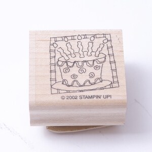 Peut inclure: Un tampon en bois avec un dessin au trait noir et blanc d'un gâteau d'anniversaire avec des bougies. Le tampon est étiqueté "© 2002 STAMPIN' UPI".