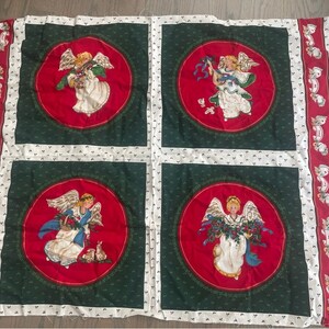 May include: A white fabric panel with four red and green squares. Each square features a circular design with a white angel holding a wreath of greenery. The angel has blonde hair and is wearing a white dress with a blue sash.