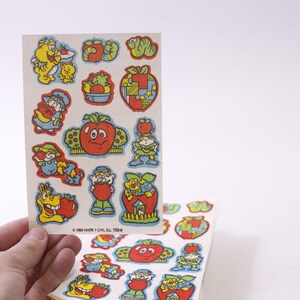 May include: Two sheets of vintage stickers with colourful cartoon characters and fruit designs. The stickers include images of smiling tomatoes, apples, and other whimsical figures. The text "© 1984 MARK I CHL, AL 75234" is printed on the bottom of the top sheet.