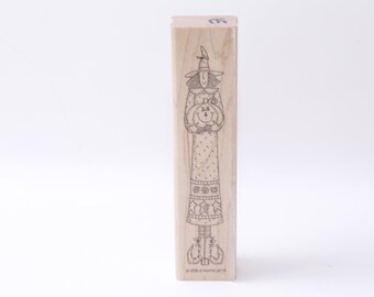 May include: A wooden stamp featuring a line drawing of a witch holding a pumpkin. The witch is wearing a pointed hat, a long dress with a patterned skirt, and boots. The text "©1999 Stampin' Up!®" is printed at the bottom of the stamp.