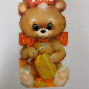 May include: A brown teddy bear wearing a red and white striped scarf and a red hat with the text "JUST FOR YOU" on it. The bear is holding a yellow jar of honey with a spoon.