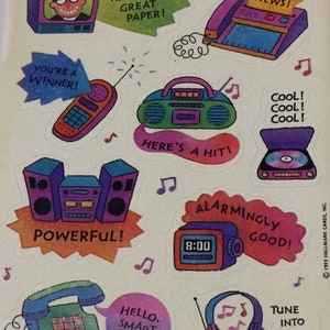 Puede incluir: Una hoja de pegatinas coloridas con un tema retro. Los diseños incluyen un televisor, una máquina de fax, un radiocasete, un teléfono, un despertador y auriculares. El texto incluye "News Flash!", "You're a Winner!" y "Hello, Smart Kid!"