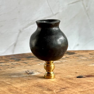 Może przedstawiać: Czarny finial do lampy z zaokrąglonym korpusem i mosiężną podstawą. Finial ma mały otwór u góry. Jest to element dekoracyjny do lamp, na jasnym tle.
