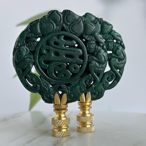 May include: Dark green jade lamp finial with intricate carvings of figures and symbols. The finial has a circular design with a central character and is mounted on two brass-colored bases. The background is blurred.