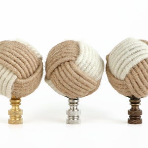 May include: Three decorative lamp finials made of woven rope in a beige and white color scheme. The finials are round and have a knot design. Each finial is attached to a metal base.
